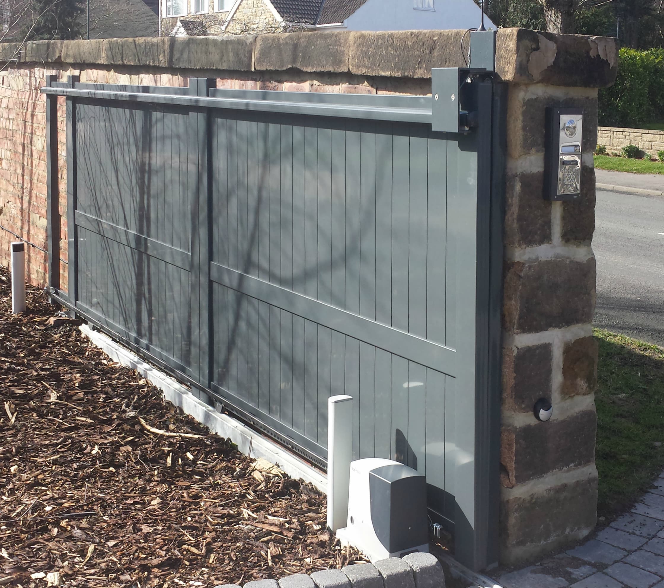 Modern aluminium sliding gate with a matte grey finish, adjacent to a stone pillar with an intercom system.